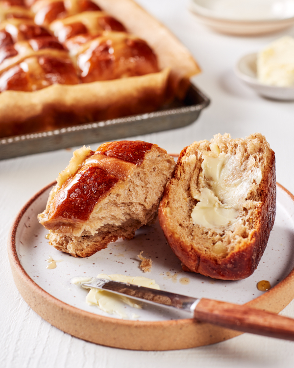 Une brioche en tranches et beurrée sur une assiette, avec une plaque de cuisson de brioches à la cuillère en arrière-plan.