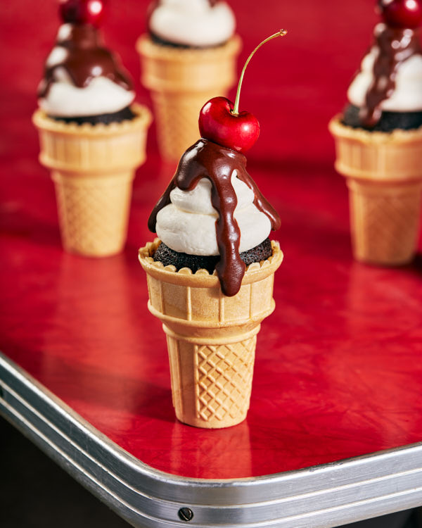 Gâteaux au chocolat dans des cornets à glace classiques, décorés de glaçage, de ganache au chocolat et d'une cerise, présentés sur une table de restaurant vintage.