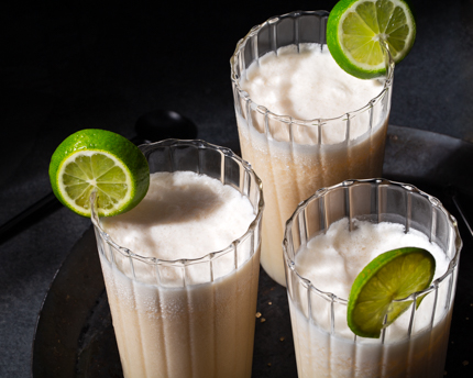 Sur une assiette noire, trois verres de kombucha à la noix de coco décorés d’une tranche de lime>