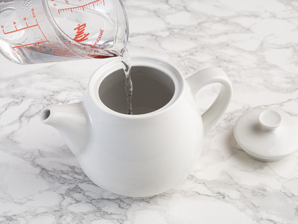 Eau bouillante qui se fait verser d’une tasse à mesurer dans une théière blanche