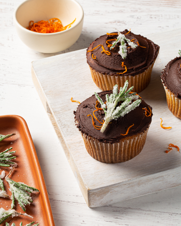 Trois cupcakes garnis de glaçage à la ganache, zeste d’orange et romarin givré sur un plateau blanchi décoré de garnitures