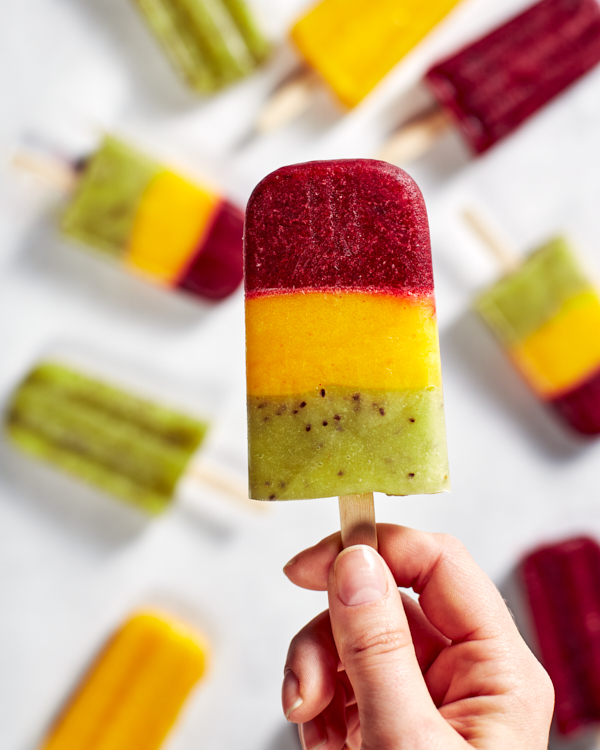 La main d'une dame tenant un paleta à trois couches de cerise, mangue et kiwi par le bâton.