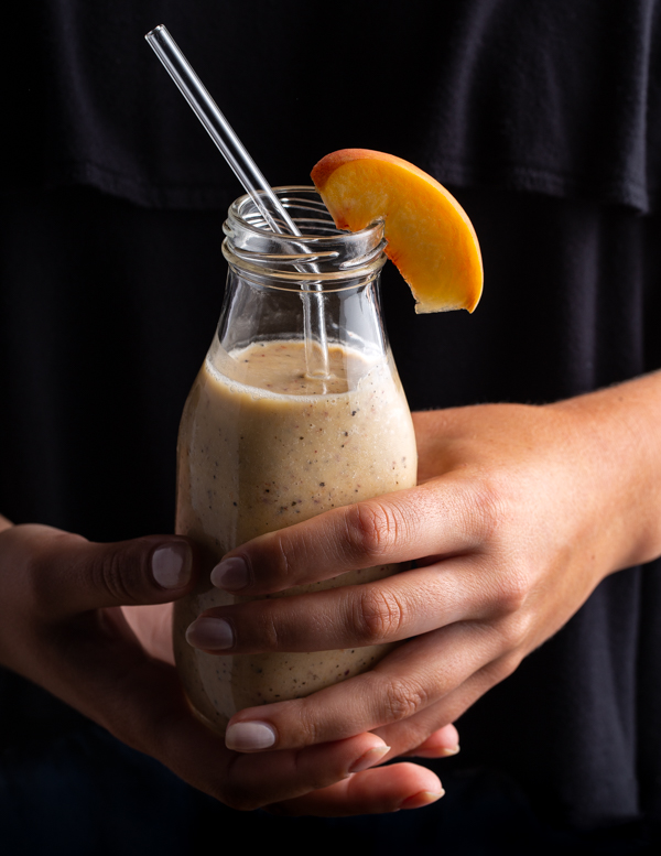 Une main féminine tenant une bouteille de portion individuelle de smoothie banane, pêche et Earl Grey avec une paille en verre, décorée d’une tranche de pêche