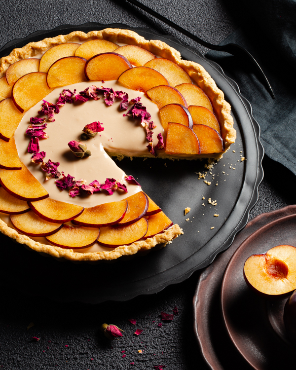 Tarte à la crème aux prunes avec une pointe manquante, décorée de tranches de prunes et de pétales de roses séchées sur un plateau noir déposé sur une table noire