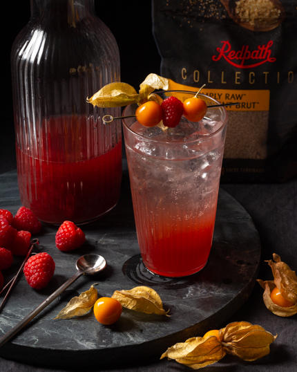 Un verre de cocktail pétillant au shrub décoré de framboises et de groseilles, disposé à côté d’une cruche de shrub framboises et groseilles, de petits fruits et d’une cuillère