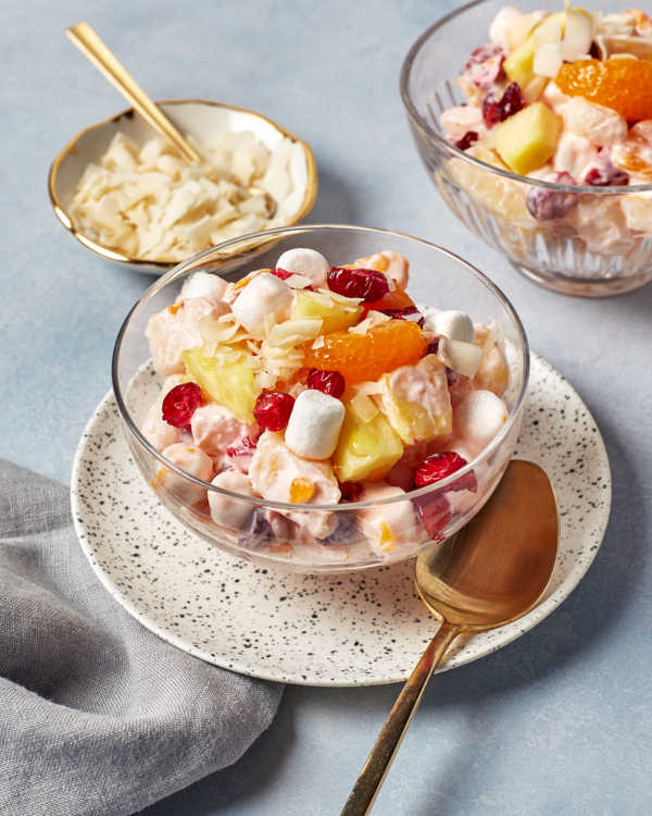 Une portion individuelle de salade d’ambroisie servie dans un bol en verre et accompagnée d’une assiette remplie de flocons de noix de coco assortie de cuillères dorées