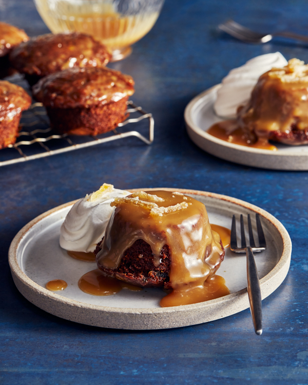 Deux sticky toffee puddings sur des assiettes, servis avec une sauce au caramel et de la crème fouettée, avec d'autres puddings sur une grille de refroidissement en arrière-plan.