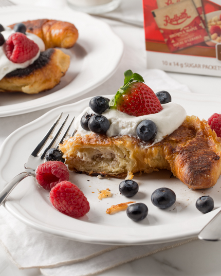 Un croissant cuit garni de crème fouettée et de petits fruits avec une bouchée manquante, et une boîte de Cassonade et Sucre d'Érable Redpath® en arrière-plan