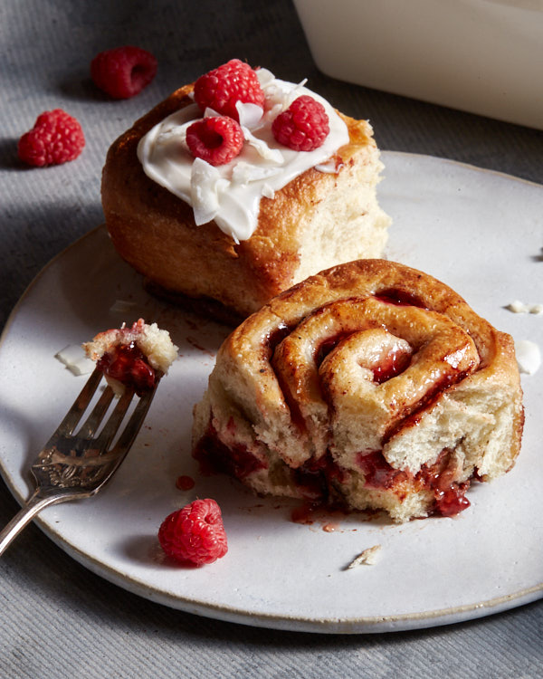 Deux roulés à la cannelle végétaliens aux framboises sur une assiette, l'un avec un glaçage à la noix de coco et des framboises, l'autre avec une bouchée manquante.