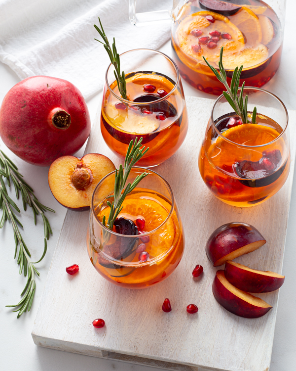 rois verres de sangria blanche décorés de glaçons, de brins de romarin et de fruits tranchés, et une grenade