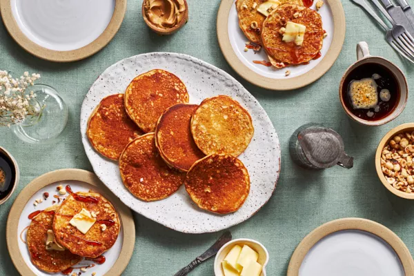 Une table dressée avec un plateau de crêpes au beurre d'arachide, du beurre, un bol de cacahuètes hachées, du beurre d'arachide, une tasse de café, des couverts, une carafe de sirop et deux assiettes avec des crêpes, du sirop et du beurre.