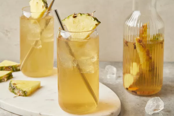 Deux verres de tepache avec des pailles en métal et garnis de quartiers d'ananas, présentés avec une carafe de tepache.