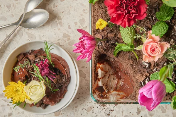 Pouding au chocolat saupoudré de miettes de biscuits et garni de fleurs comestibles dans un plat à four en verre, présenté avec une portion de pudding dans un bol et des cuillères.