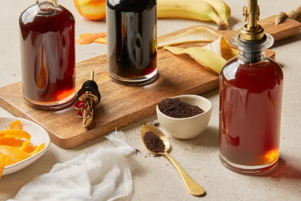 Une bouteille de sirop simple au café, une bouteille de sirop simple à l'orange et une bouteille de sirop simple à la banane sont présentées sur une table avec une planche de service, accompagnées de marc de café, de pelures d'orange et de pelures de banane.