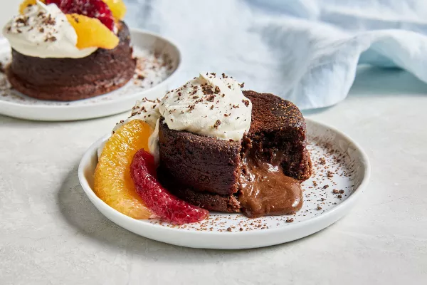 Deux gâteaux individuels au chocolat fondant garnis de crème fouettée et de cacao en poudre, agrémentés de segments d'orange et d'orange sanguine. L'un des gâteaux est montré avec un morceau manquant, laissant échapper une sauce au chocolat à partir du centre