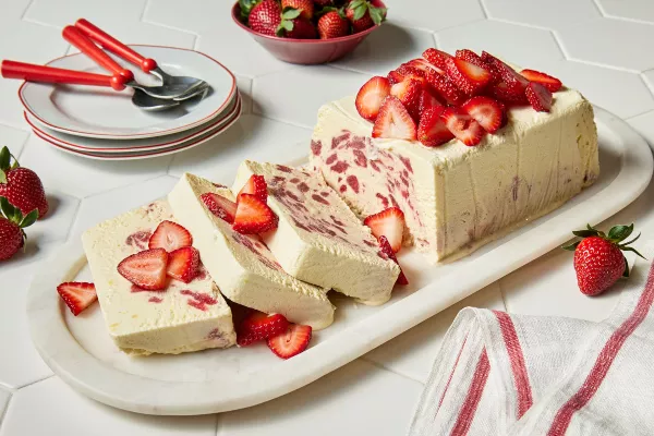 Un pain rectangulaire de semifreddo à la fraise et à la fleur de sureau sur un plateau en marbre, garni de tranches de fraises, avec trois tranches coupées, présenté avec trois assiettes blanches à bordure rouge, trois cuillères rouges et un bol rouge de fraises.