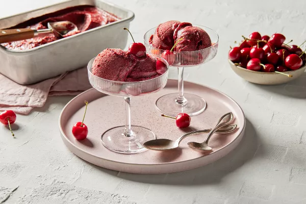 Deux coupes à pied de sorbet aux cerises garnies de cerises, sur un plateau rose pâle avec des cuillères en or, présentées avec un bol de cerises et une casserole de sorbet aux cerises avec une cuillère à boule pour servir.