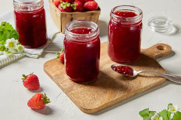 Trois pots de confiture de fraises, deux sur une planche à découper en bois et un sur un torchon, présentés avec une cuillère de confiture, des fraises fraîches et des fleurs de fraises.