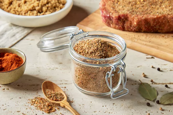 Un bocal en verre avec un couvercle rempli d'épices sucrées pour steak à la montréalaise est présenté avec un steak cru assaisonné sur une planche à découper, un bol de sucre brun et un bol d'épices. 