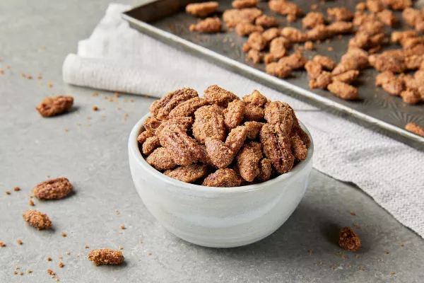 Un bol blanc débordant de pacanes et d’amandes confites, déposé sur un comptoir de cuisine avec une plaque à pâtisserie de noix préparées.