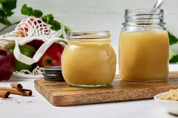 Deux bocaux en verre de compote de pommes maison sur une planche à découper en bois sur une surface blanche, un sac en maille de pommes, des bâtons de cannelle et un bol de Cassonade Dorée.