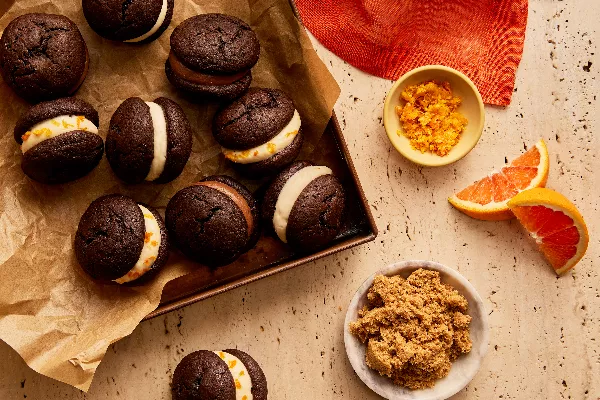 Des mini whoopie pies au chocolat avec diverses garnitures sur une plaque de cuisson doublée de papier parchemin, présentés avec un bol de sucre brun foncé, un bol de zeste d'orange et des quartiers d'orange.