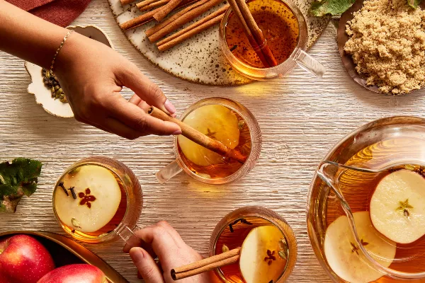 Quatre tasses en verre de cidre de pomme chaud garnies de tranches de pomme, de clous de girofle et de bâtonnets de cannelle, présentées sur une table avec une carafe de cidre, un bol de pommes et un bol de sucre jaune doré, et des mains tendues vers les tasses.