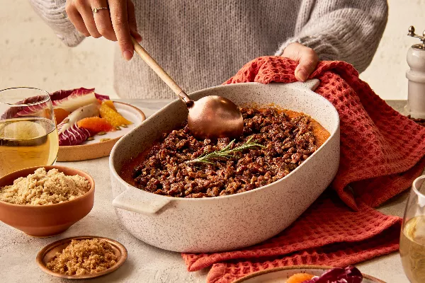 Une femme portant un chandail sert une casserole de patates douces garnie d'une branche de romarin depuis un plat de cuisson, présentée avec une salade de radicchio et d'agrumes, des verres de vin et des bols de sucre jaune doré et de sucre Demerara.