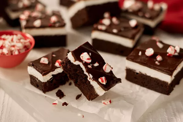 Un gros plan de brownies au chocolat menthe avec une couche de crème à la menthe et un nappage de ganache, chacun saupoudré de morceaux de canne en bonbon, présentés sur du papier ciré sur une surface blanche.