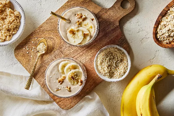  Vue de dessus de deux grands verres de smoothie à la banane, avoine et tahini, présentés sur une planche à découper en bois avec des bols de sucre jaune doré, de graines de sésame et d'avoine sèche, accompagnés de deux bananes non pelées.