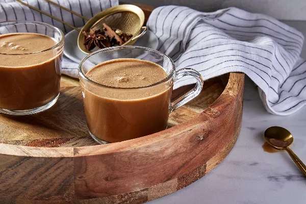 Deux tasses en verre de masala chai sur un plateau de service rond en bois, accompagnées d'une passoire dorée contenant des épices entières, d'un torchon et d'une cuillère dorée.