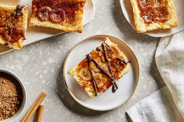 Quatre tartes renversées à la banane, deux dans des assiettes et deux sur un plateau, présentées avec un bol de sucre brun et des bâtonnets de cannelle.