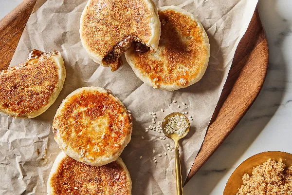 Cinq crêpes coréennes ou hotteok sur un plateau en bois, deux ouvertes avec du sirop de sésame qui s'écoule, présentées avec des graines de sésame éparpillées et un bol de cassonade de style Demerara.