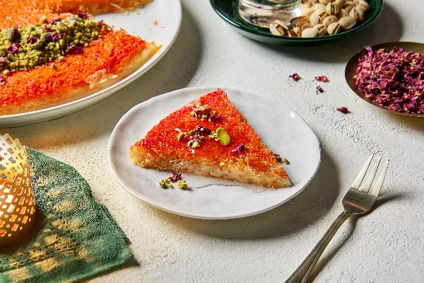 Une tranche de knafeh dans une assiette, garnie de pistaches et de pétales de fleurs comestibles, présentée avec un knafeh entier, des plats de pistaches en coque et de pétales de fleurs, et une bougie allumée.