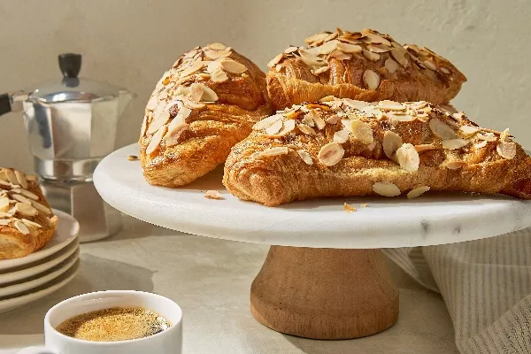 Quatre croissants aux amandes cuits à la friteuse à air, garnis d'amandes tranchées sur un présentoir à gâteaux blanc, avec une tasse d'espresso et une cafetière à l'arrière-plan.