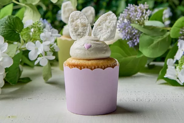  Cupcakes au citron décoratifs sur le thème de Pâques avec un glaçage à la crème au beurre et des oreilles de lapin en guimauve trempées dans la noix de coco, présentés dans des caissettes à cupcakes pastel, parfaits pour un dessert de printemps festif.