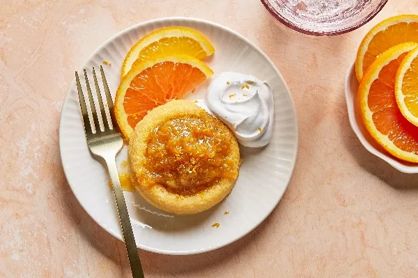 Vue en plongée d'un gâteau éponge unique nappé de marmelade d'agrumes brillante et de zeste, servi sur une assiette blanche cannelée avec des tranches d'orange fraîches et une touche de crème fouettée, accompagné d'une fourchette, sur un fond aux tons pêche doux.