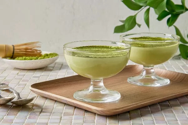 Deux tasses en verre de mousse au chocolat blanc et matcha garnies de poudre de matcha verte sur un plateau en bois, avec un fouet à matcha traditionnel et un bol de poudre de matcha en arrière-plan, posés sur une table carrelée à la lumière naturelle.