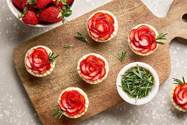 Cinq mini-tartelettes fraises et romarin sur une planche à découper en bois, présentées avec un bol de brins de romarin, un bol de fraises fraîches et une sixième mini-tartelette déposée sur le comptoir.