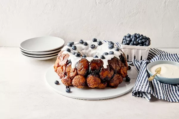 Monkey bread estival aux bleuets sur une assiette blanche avec du glaçage blanc coulant et des bleuets frais parsemés sur le dessus, disposé à côté d’une serviette rayée bleue et blanche, d’un bol bleu contenant du glaçage et d’un panier de bleuets, le tout sur une surface de couleur pâle.