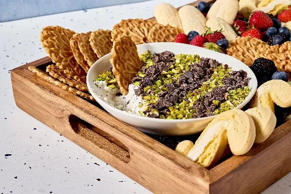 Un plateau en bois rempli d'un assortiment de biscuits italiens et de fruits frais - des fraises, des bleuets et des mûres - entoure un bol de trempette à cannoli. La trempette est garnie de pistaches hachées et de morceaux de chocolat, et on y voit un biscuit Pizzelle en train de tremper.