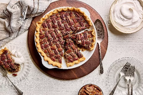 Une tarte au pacane dans un plat à gratin, montrant trois tranches coupées, dont une servie sur une assiette avec de la crème fouettée, sur une table en bois blanc avec un bol de crème fouettée et un bol de pacanes.