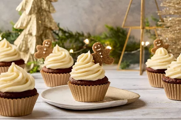 Des petits gâteaux au pain d'épices festifs surmontés d'un tourbillon de glaçage blanc crémeux et d'un mini biscuit au pain d'épices. Les petits gâteaux sont disposés sur une surface de bois avec des décorations de Noël en arrière-plan, dont un sapin de Noël doré et des lumières féériques scintillantes.