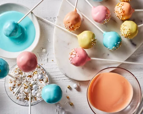 Des cake pops aux couleurs pastel avec des éclats de verre sur un plateau, certains dans un verre d'éclats de verre et un autre 