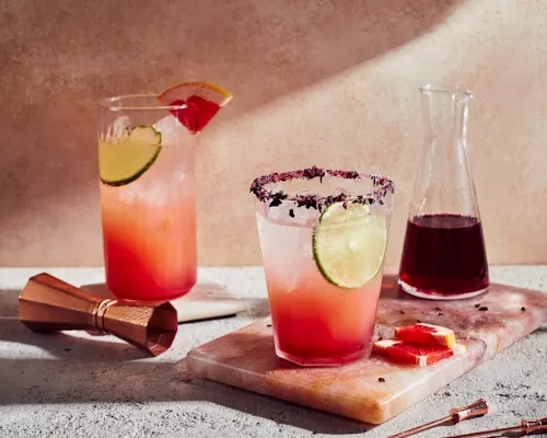 Deux verres de paloma à l'hibiscus avec un pichet de sirop simple, présentés avec un jigger et des brochettes.