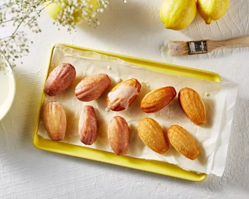 Des madeleines au citron glacées sur un plateau jaune avec un bol de glaçage, un pinceau à pâtisserie et des citrons.