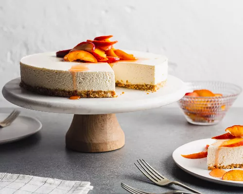 Gâteau au fromage à la vanille sans cuisson avec pêches macérées sur une assiette à gâteau, et un morceau de gâteau dans une ass