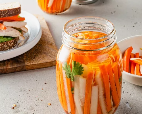 Un bocal de carottes et de radis daikon marinés avec un sandwich en arrière-plan.