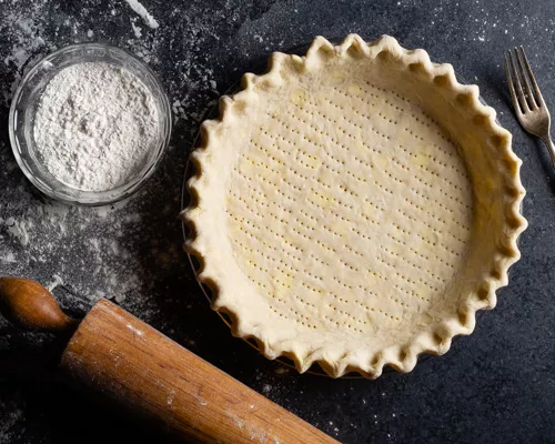 Une croûte à tarte non cuite avec un bol de farine, un rouleau à pâtisserie et une fourchette