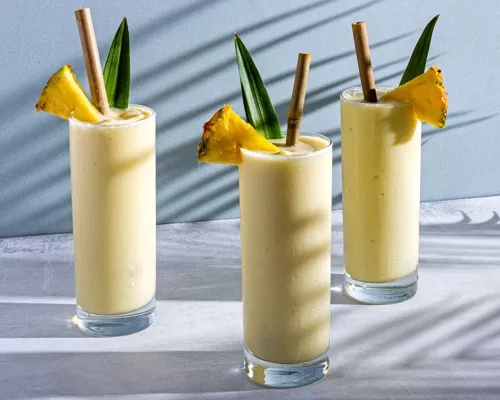 Trois verres de pina colada garnis de bâtonnets et de feuilles d’ananas avec des pailles en bambou